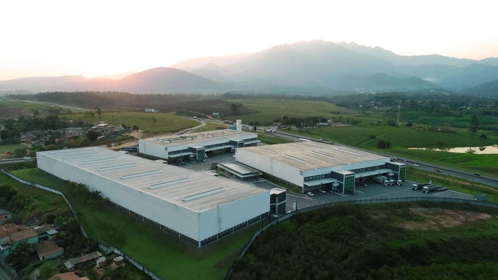 Condomínio logístico ao entardecer, às margens de rodovia, com serra ao fundo